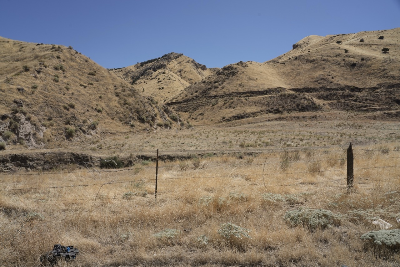 An eye-level photograph of a dry, golden-brown hillside featuring weathered wooden fence posts, rusted barbed wire, sparse vegetation, and rolling dry hills under a clear blue sky.