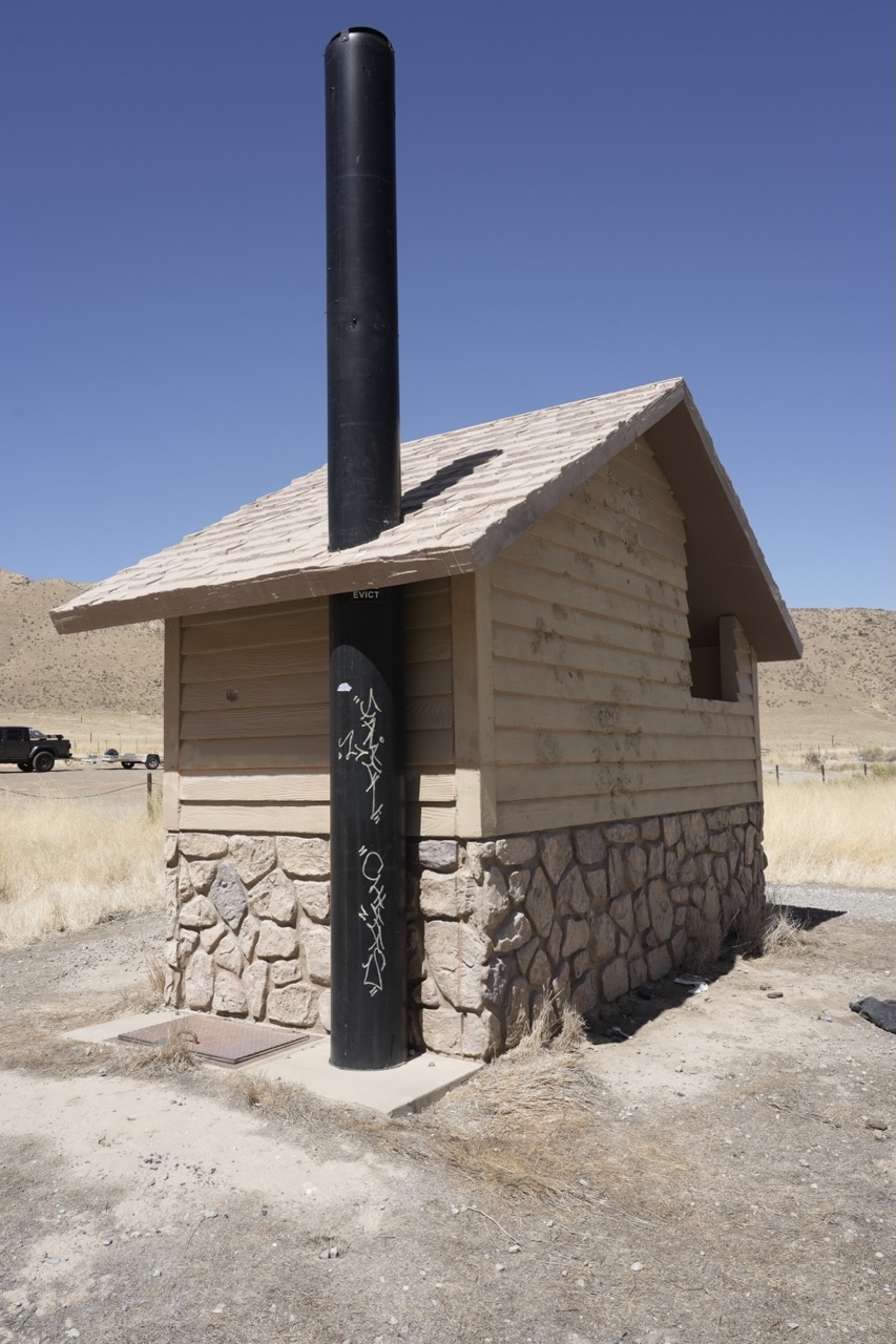 An eye-level photograph of a small, single-story utility or restroom building with a stone base, wood-paneled siding, a shingled roof, and a tall black vent pipe running up the front wall, which features white graffiti markings.
