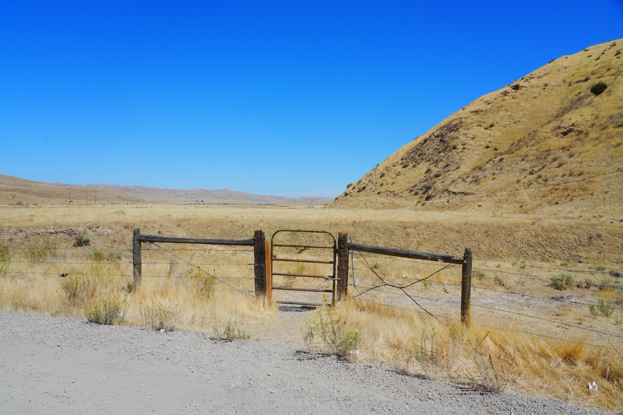 A wide-angle landscape photograph showing a rustic wooden fence with a small metal gate alongside a gravel road, opening up to a flat valley flanked by low, arid hills under a blue sky.