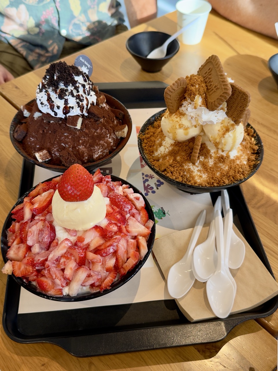 Three overflowing bowls of Korean shaved ice dessert (bingsu) arranged on a black tray: fresh strawberry topped with ice cream, chocolate with brownie bites and whipped cream, and caramel Biscoff cookie crumble.