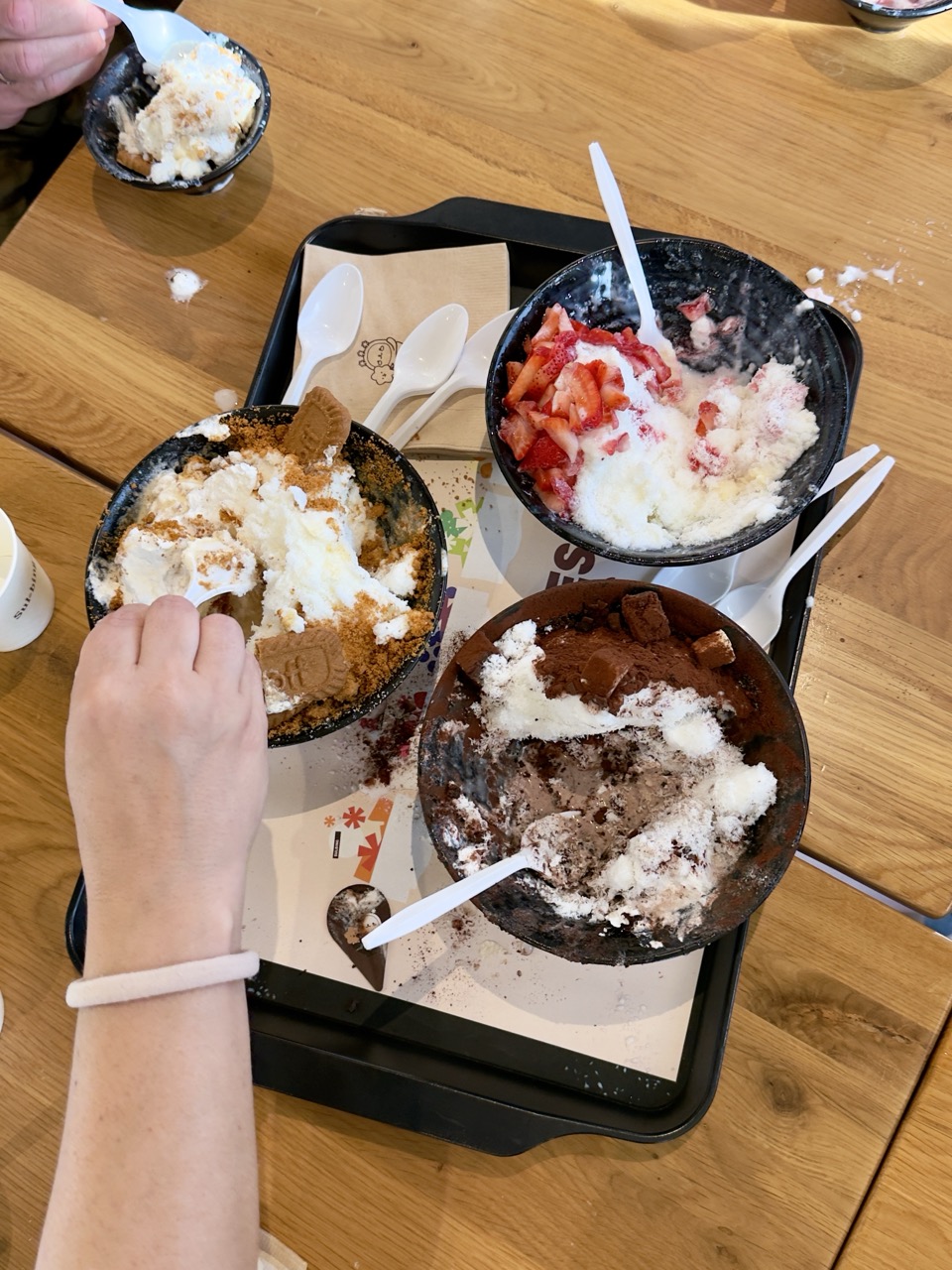Top-down view of three partially eaten bowls of bingsu on a black tray, featuring chocolate, strawberry, and Biscoff flavors, with a hand reaching in with a spoon.