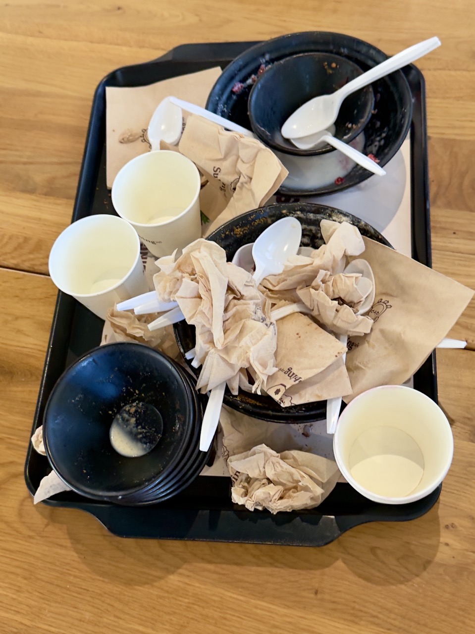 Overhead view of a black serving tray covered in finished dessert bowls, crumpled brown napkins, empty paper cups, and discarded plastic spoons on a wooden table.