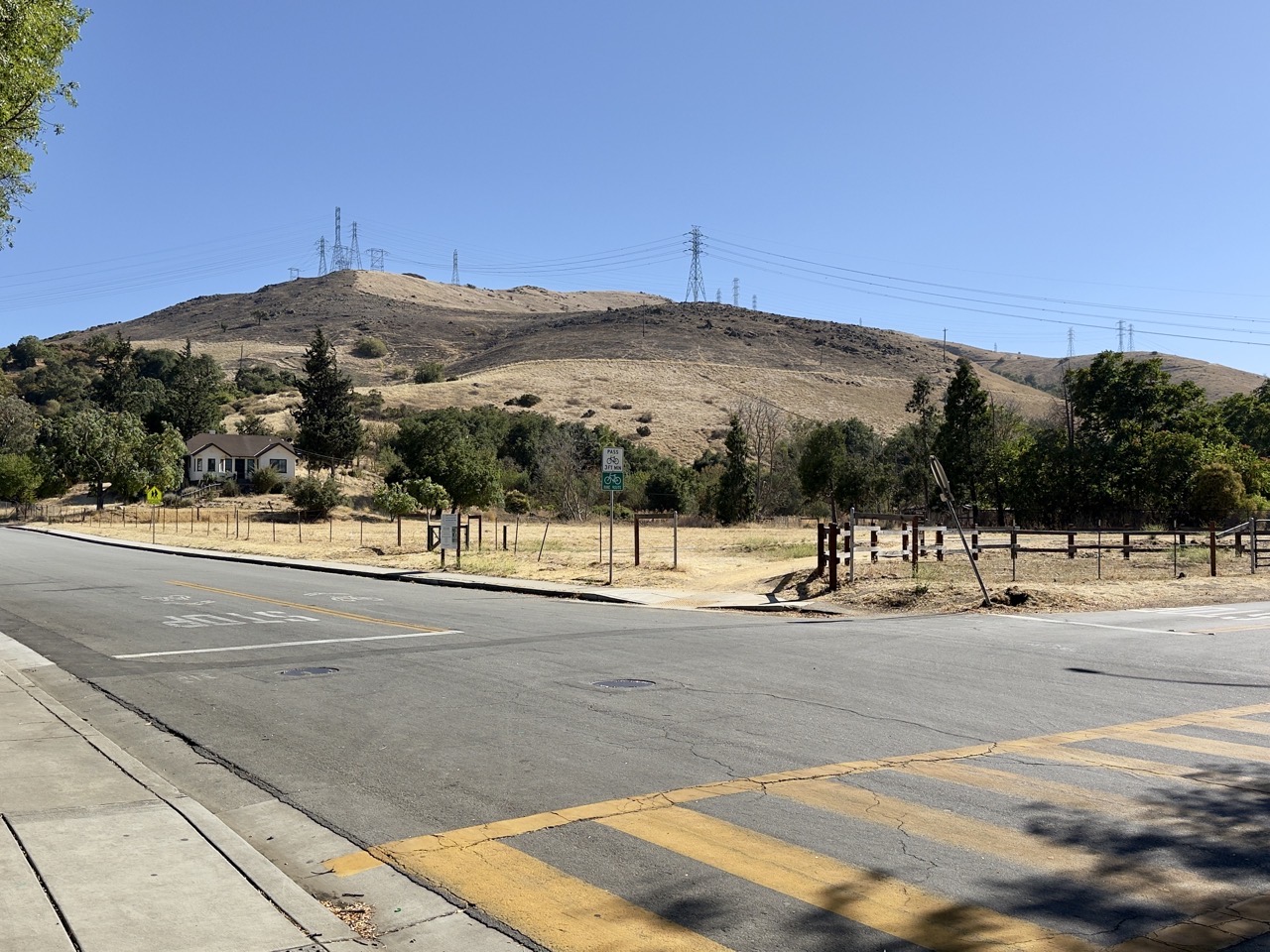 An asphalt road and yellow crosswalk in front of dry grassy hills topped with high-voltage power lines under a clear sky.