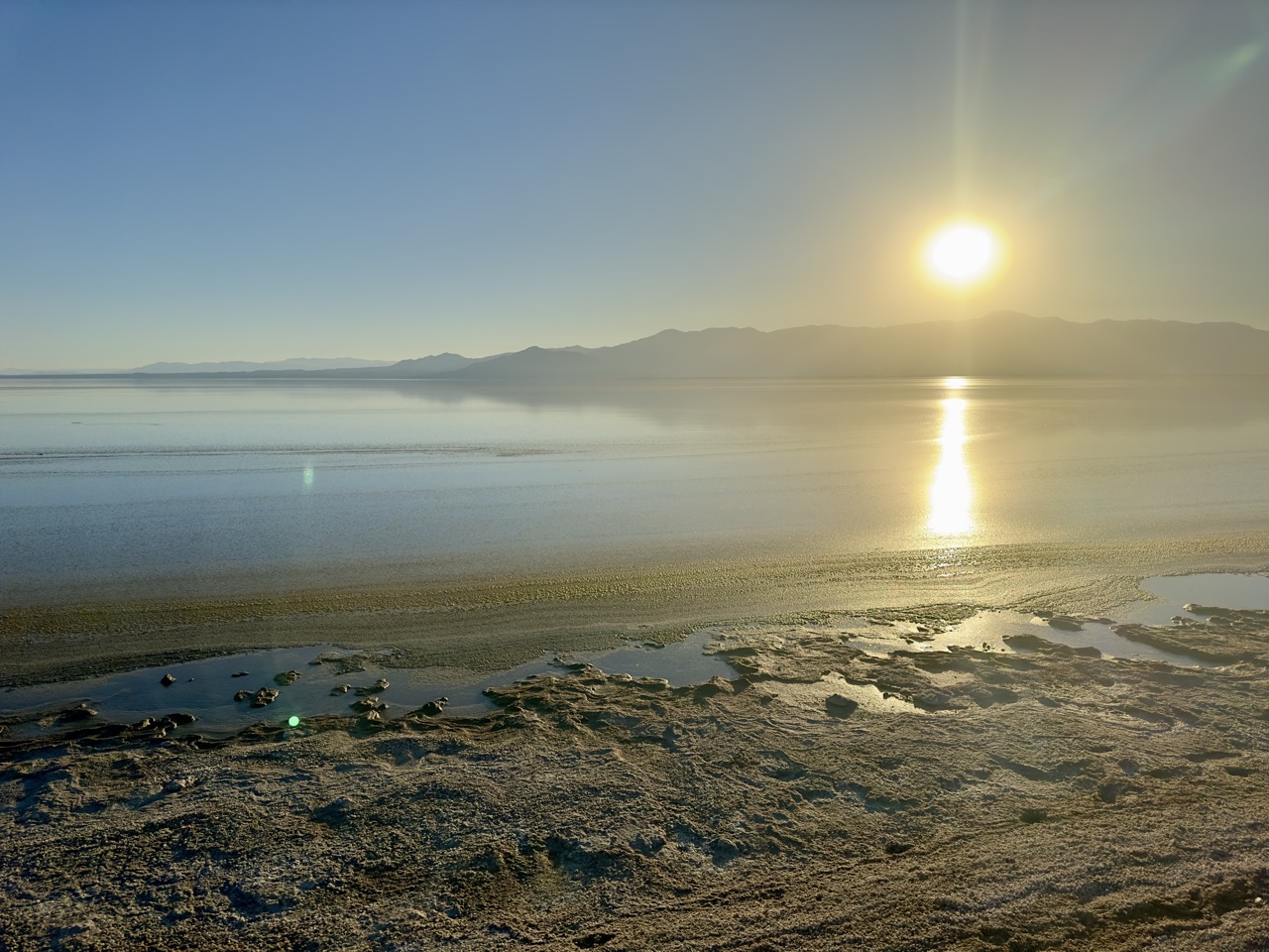 The sun low over a flat, mineral-crusted shoreline, its reflection stretching across the still water.