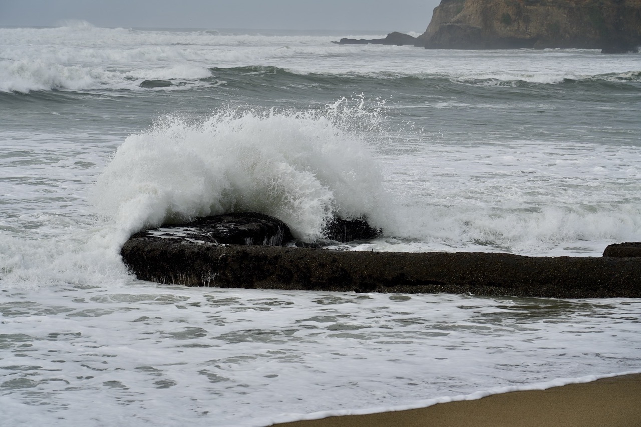 A wave bursting white over a low flat rock shelf at the shoreline, headlands standing in the haze beyond.