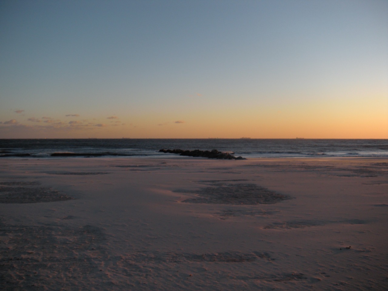A wide beach patched with snow at sunset, a low rock jetty at the waterline and freighters strung along the horizon.