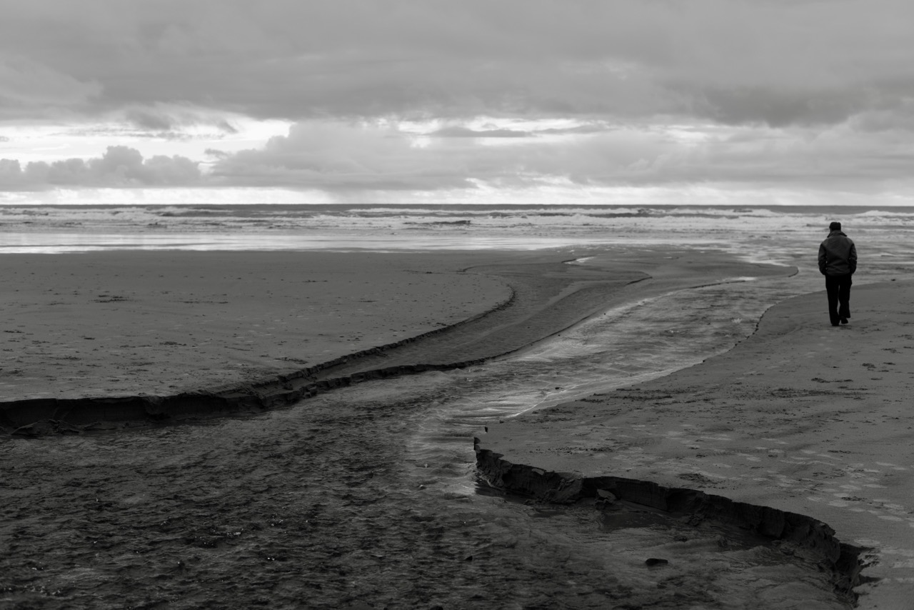 Black and white: a stream cuts a winding channel across a wide flat beach toward the surf, a lone figure in a coat walking near the water’s edge.