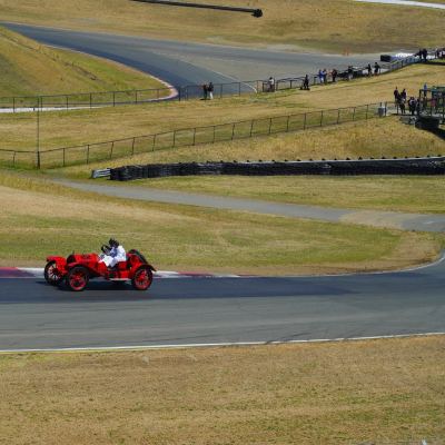 A red brass-era open-wheel racer on the track, the driver in white, dry golden hills behind.