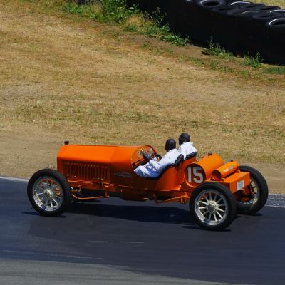 An orange two-seat vintage racer, number 15, carrying a driver and passenger past a tire wall.
