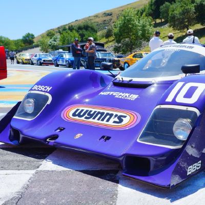 A purple Wynn's-liveried Porsche prototype, number 10, parked on a checkered paddock, dry hills behind.