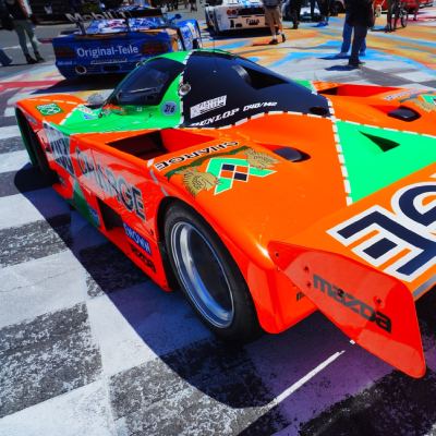 The rear of an orange-and-green Mazda 'Charge' Le Mans prototype on a painted paddock floor, a Ferrari F40 beyond.