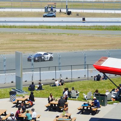 Spectators at picnic tables on a shaded patio as a car streaks past behind the catch fence.