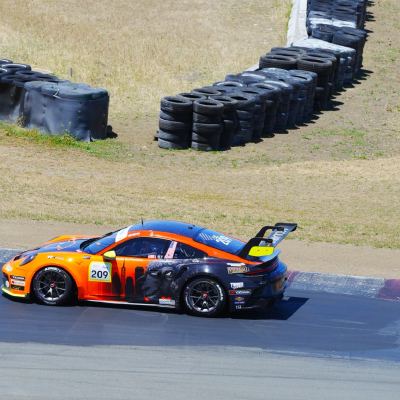 An orange-and-black Porsche 911 cup car, number 209, turning in below stacked tire barriers on the hillside.