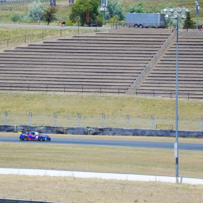 A blue-and-red car on a far stretch of track below empty grandstand terraces on the dry hillside.