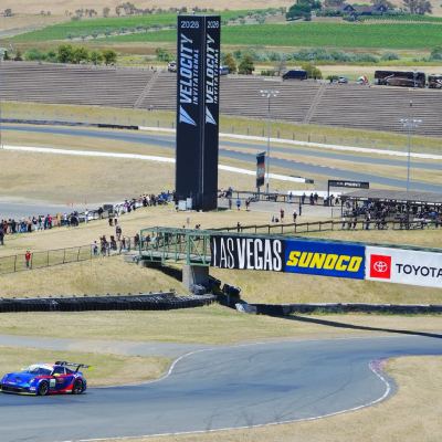 A blue-and-red Porsche GT car on the track beneath the Velocity banner tower, vineyards on the hills beyond.