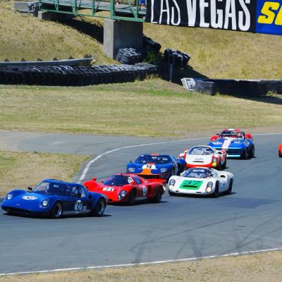 A pack of 1960s sports-racers rounding a turn, a dark-blue car and a red car leading, trackside banners behind.