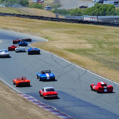A field of vintage sports cars strung down a curving descent, Cobras, coupes and roadsters among them.