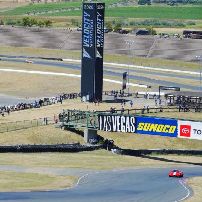 A wide view of the circuit, a blue and a red car descending toward a turn, the Velocity tower, vineyards and empty grandstands behind.