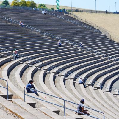Empty concrete grandstand terraces with a scattering of spectators in the sun.