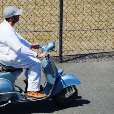 A man in white riding a light-blue vintage Vespa scooter past a fence, a microphone strapped to a wooden crate on the back.