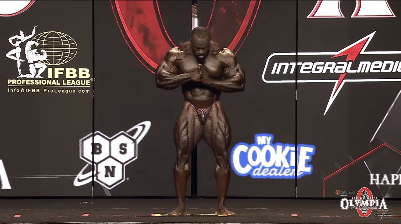 Professional bodybuilder Samson Dauda stands center stage at the 2026 IFBB Mr. Olympia competition with his head bowed and hands pressed together in front of his chest, flexing his shoulders, arms, and quadriceps before a black sponsor backdrop.