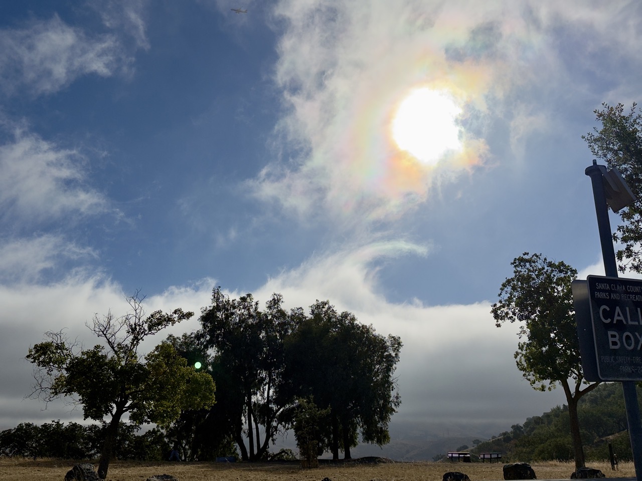 The sun breaking through wispy, torn clouds over a hilltop park, with silhouetted oak trees and a county-park call-box sign in the foreground.