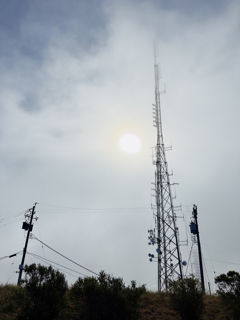 A tall three-sided lattice radio tower studded with reflector dishes and stubby antennas, silhouetted against a hazy sky with the sun behind it and a power-line pole alongside.